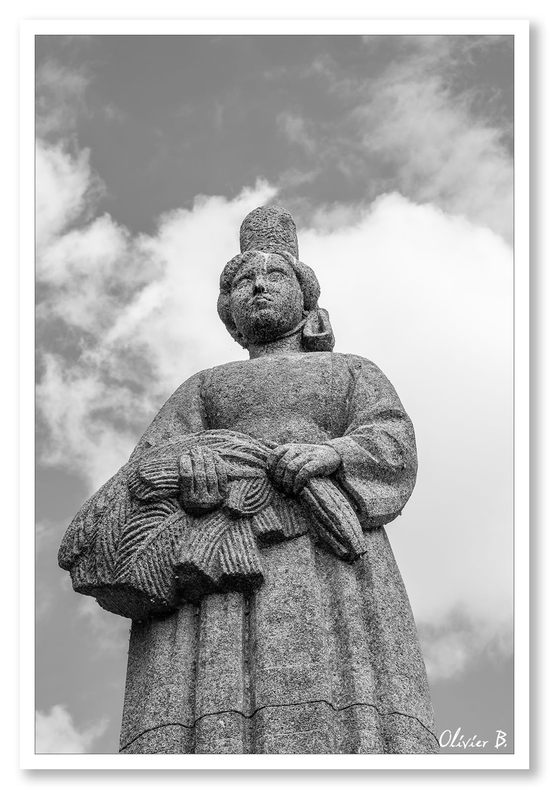 Statue en granit d'un Bigouden en contre-plongée, défiant le ciel nuageux